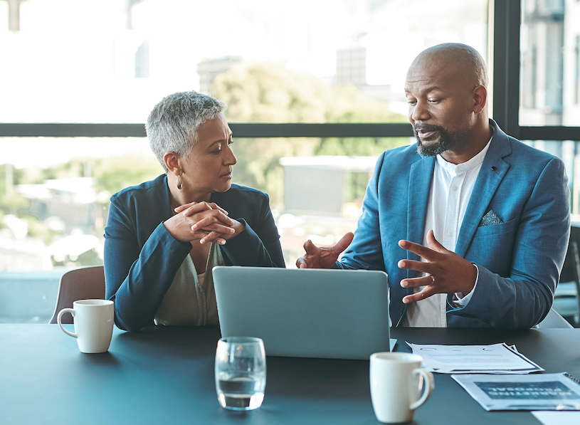A woman and a man talk in front of a laptop computer on a conference table.